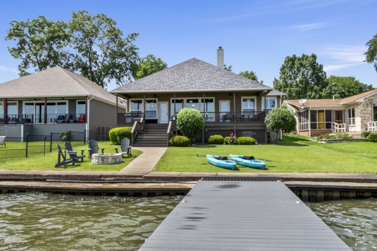 From the dock, take in the full lakeside retreat—Adirondack chairs cozying up around the fireplace, two kayaks ready for adventure, a spacious porch, and a large, lush green yard.